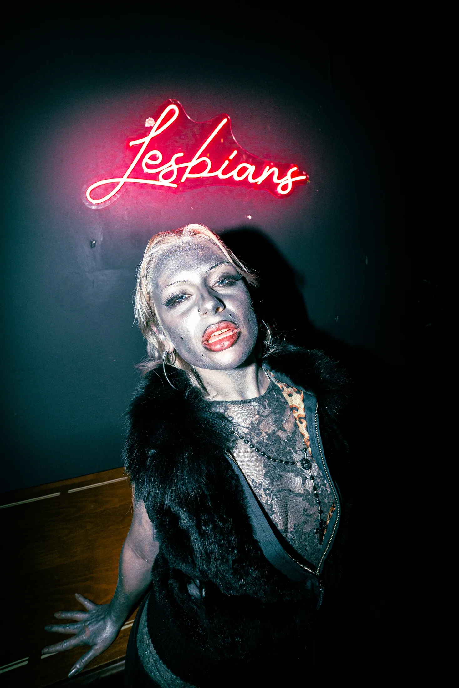 A person in a black faux-fur vest poses beneath a red neon sign.
