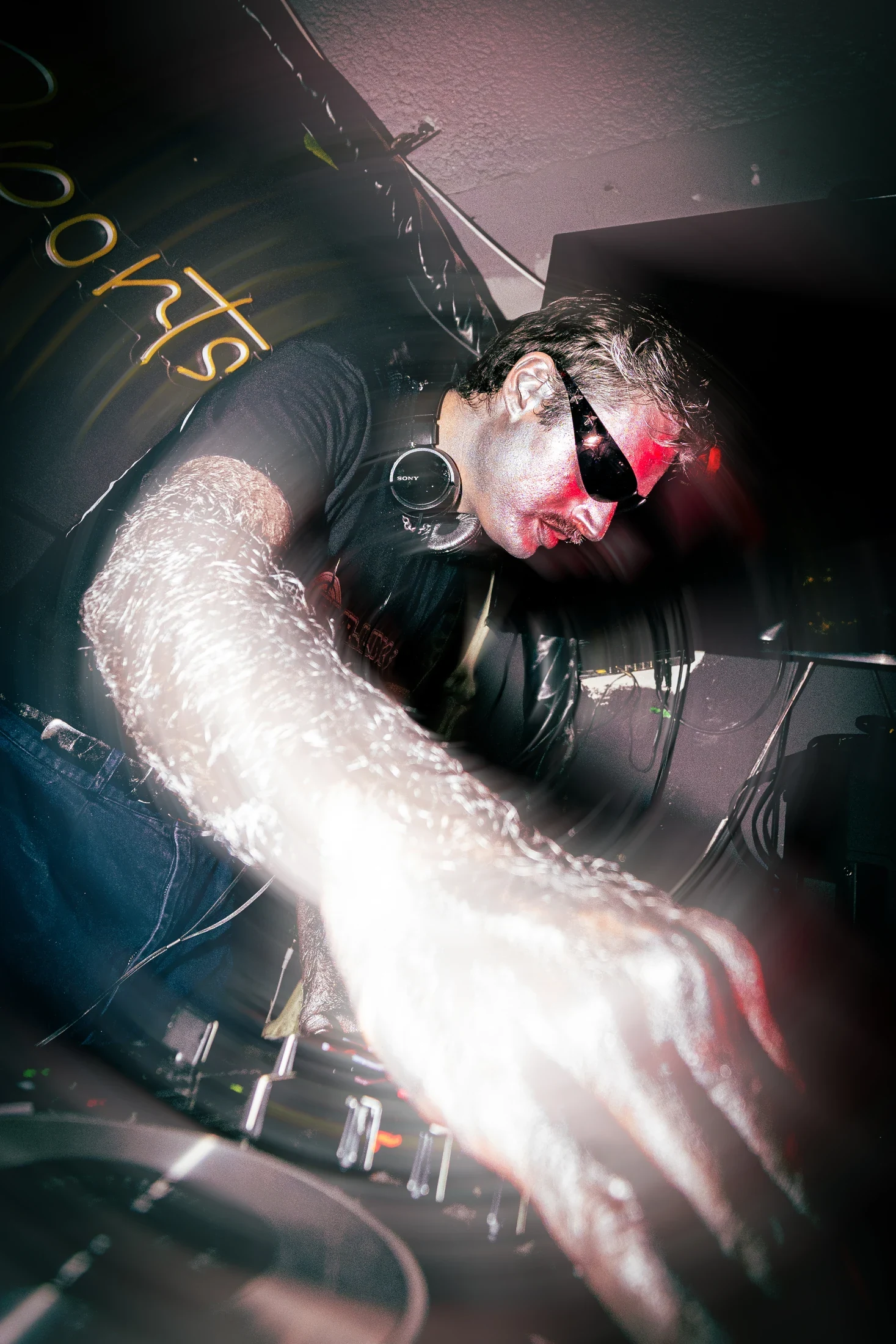 A DJ in sunglasses reaches across the decks, with motion trails around the outstretched arm.