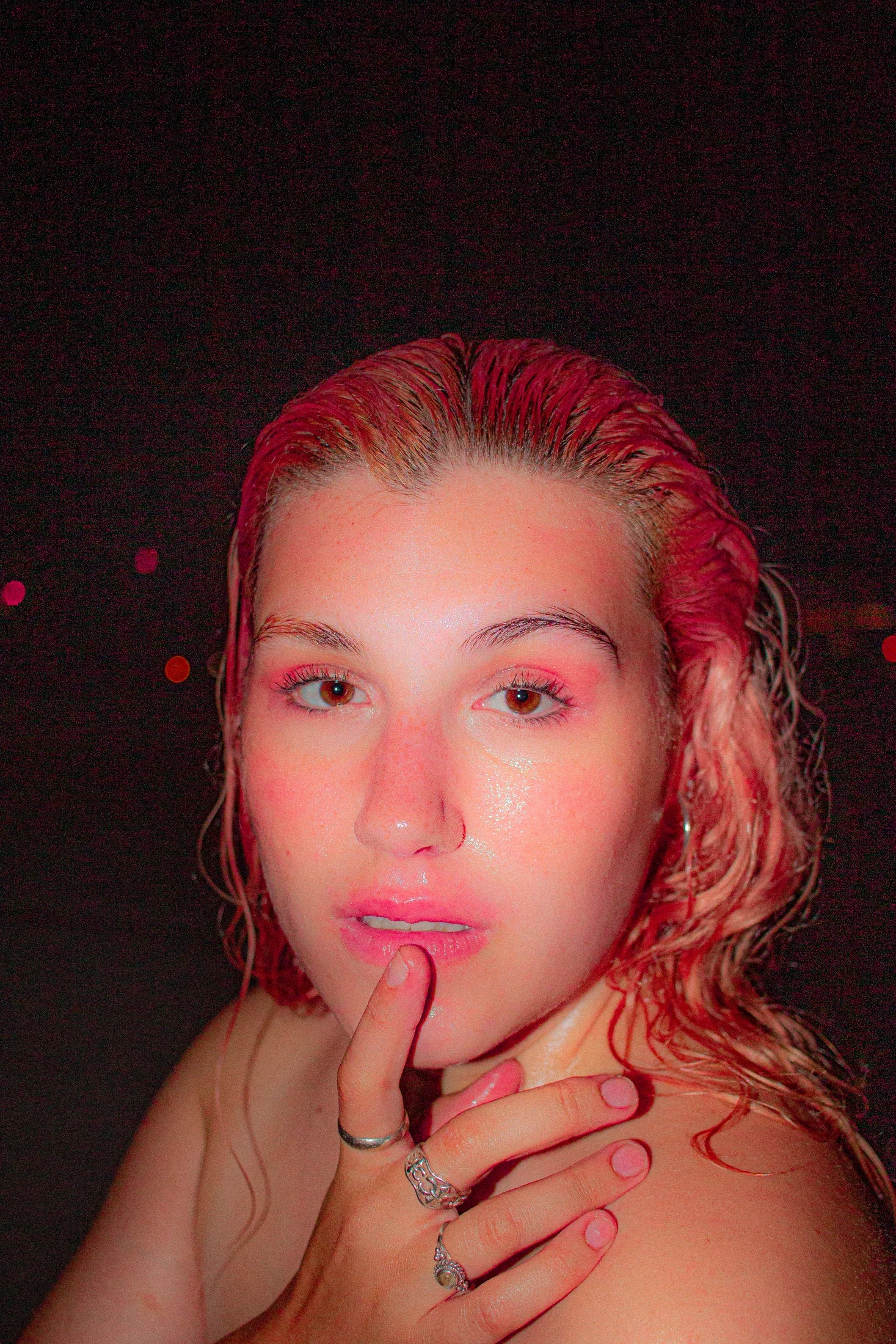 Close portrait with slicked-back pink hair, glossy makeup, and a ringed hand resting at the lips.