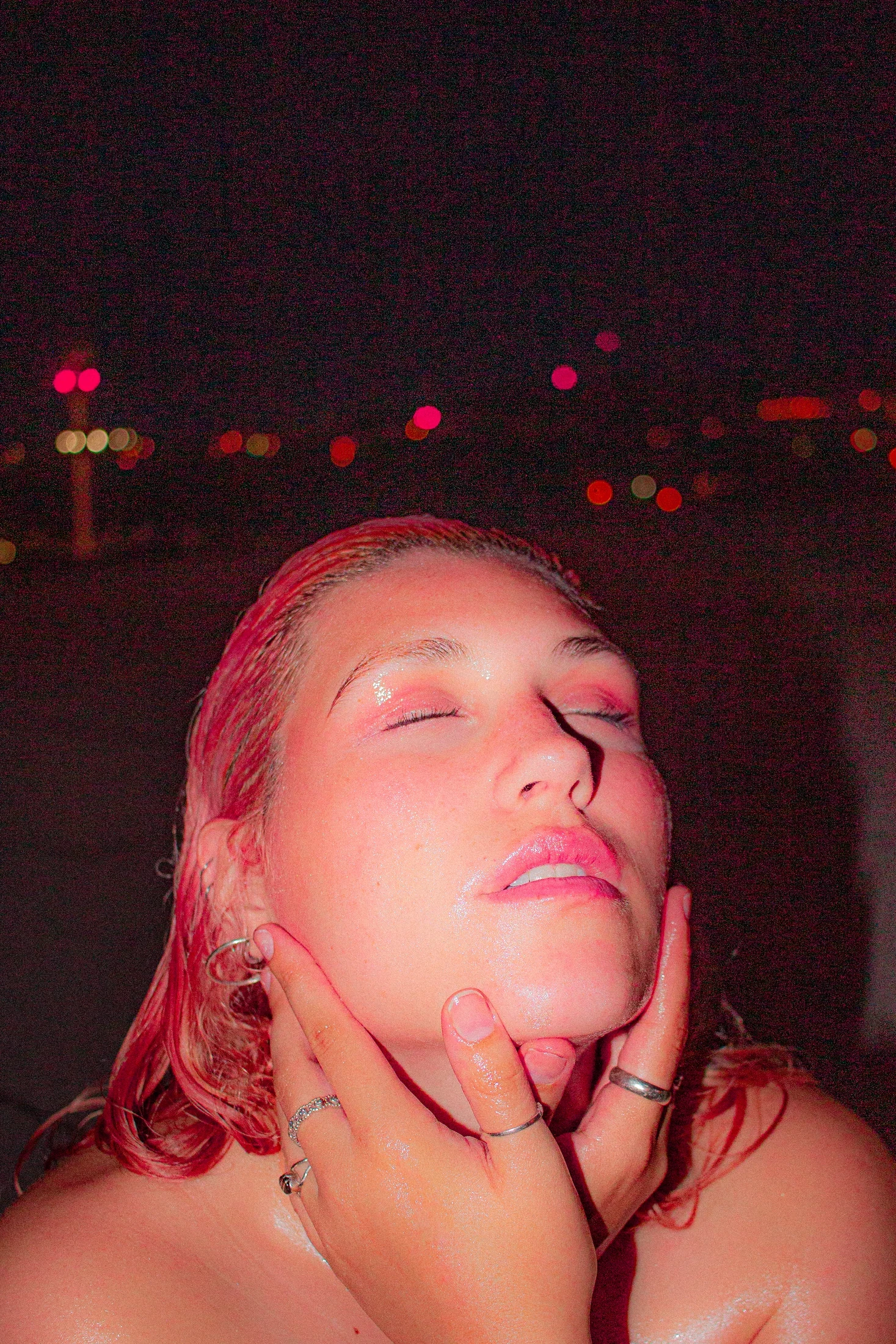 A portrait with closed eyes and hands framing the face, against distant coloured night lights.