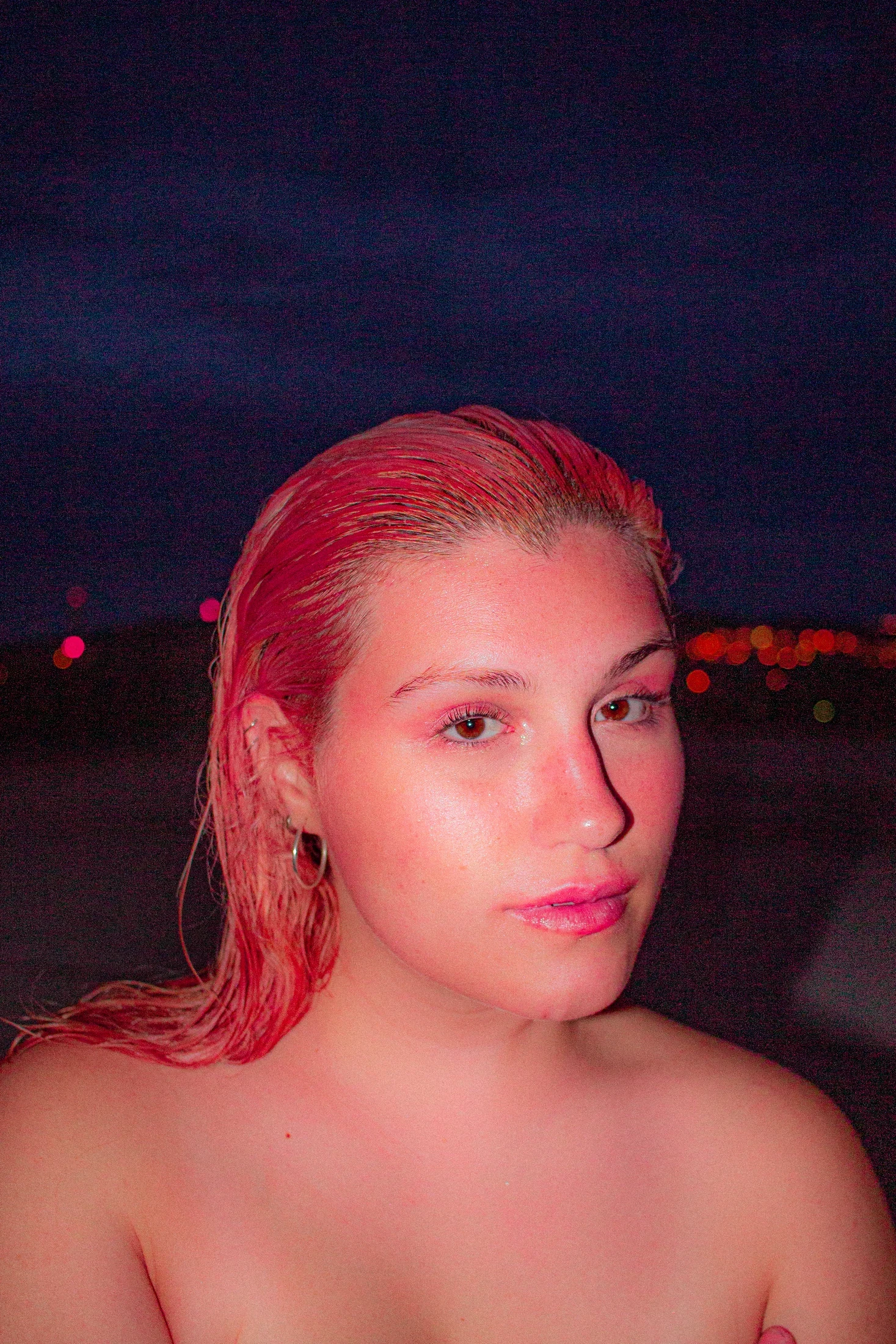 A direct-flash portrait with pink hair and makeup against a dark blue evening sky.