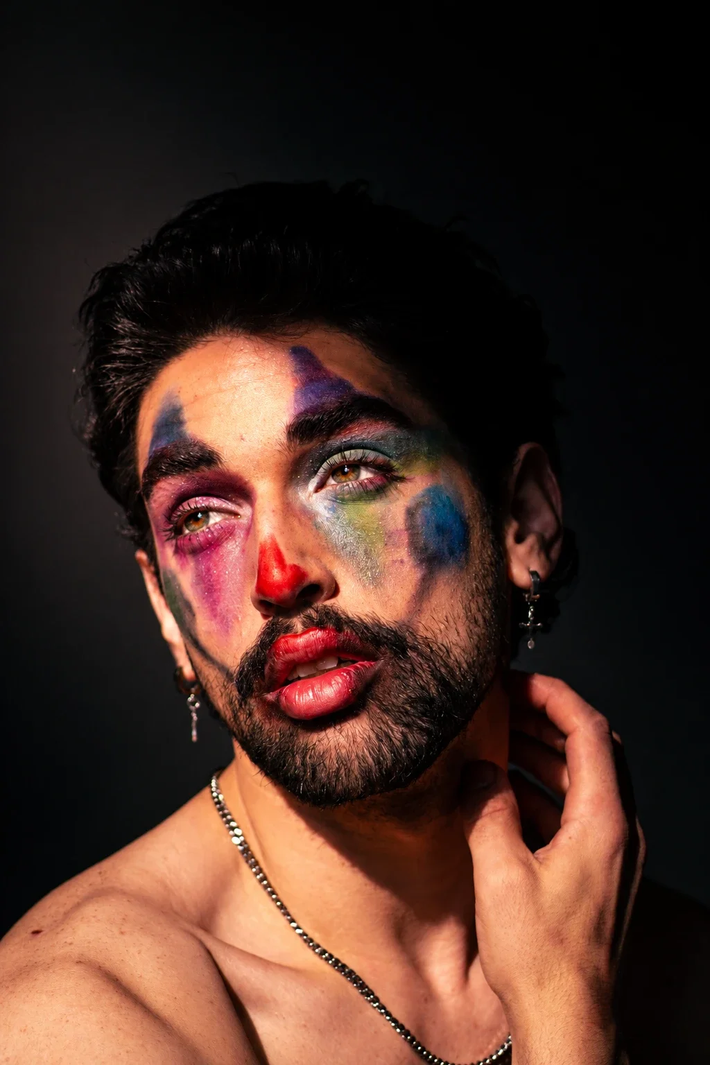 A person with colourful clown-inspired makeup looks upward, one hand resting against the neck.