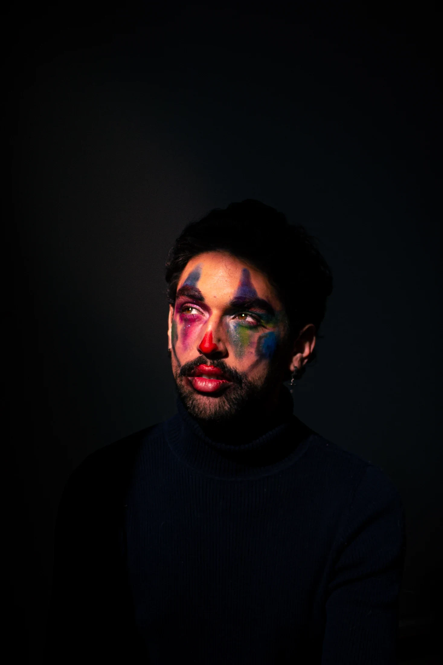 A person in a dark turtleneck and colourful face paint sits against a dark background.