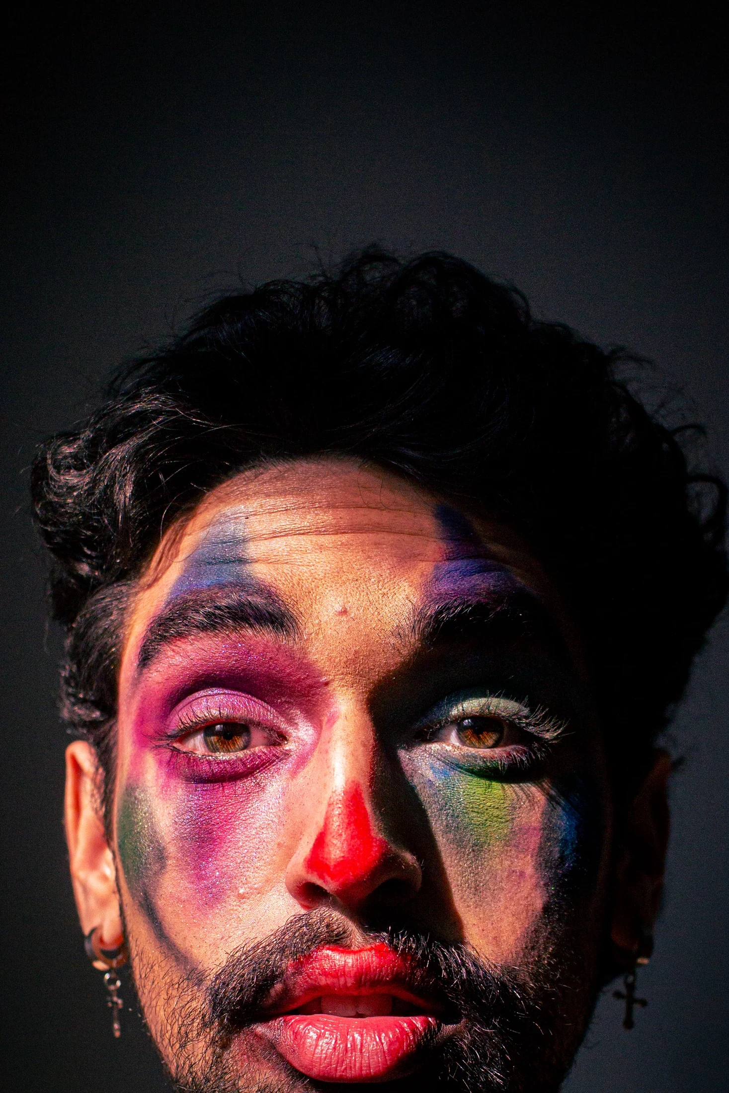 Tight portrait showing multicoloured makeup, red lips, and a direct gaze in hard directional light.
