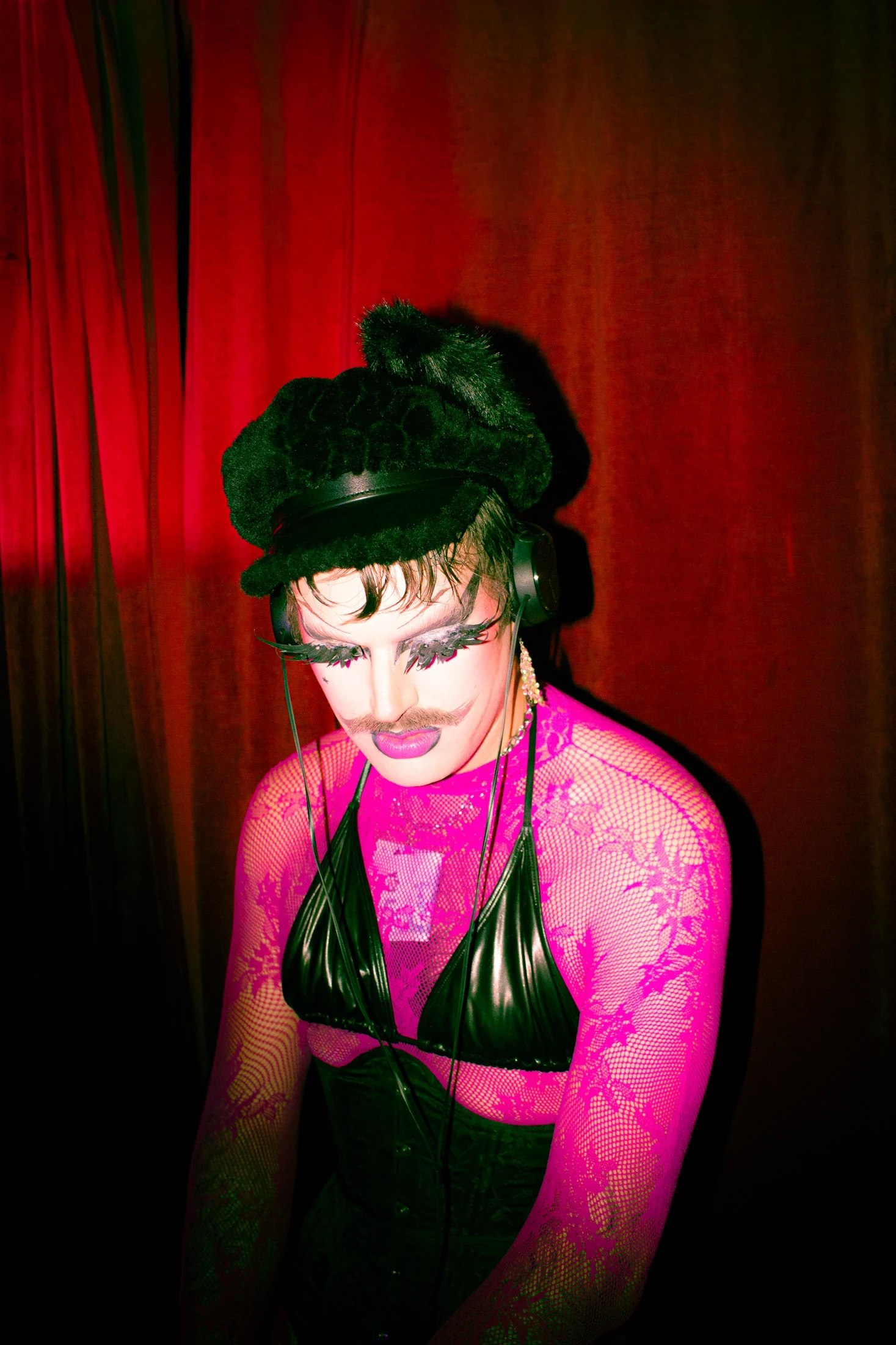 A performer in pink lace and a black corset looks into the camera against a red curtain.