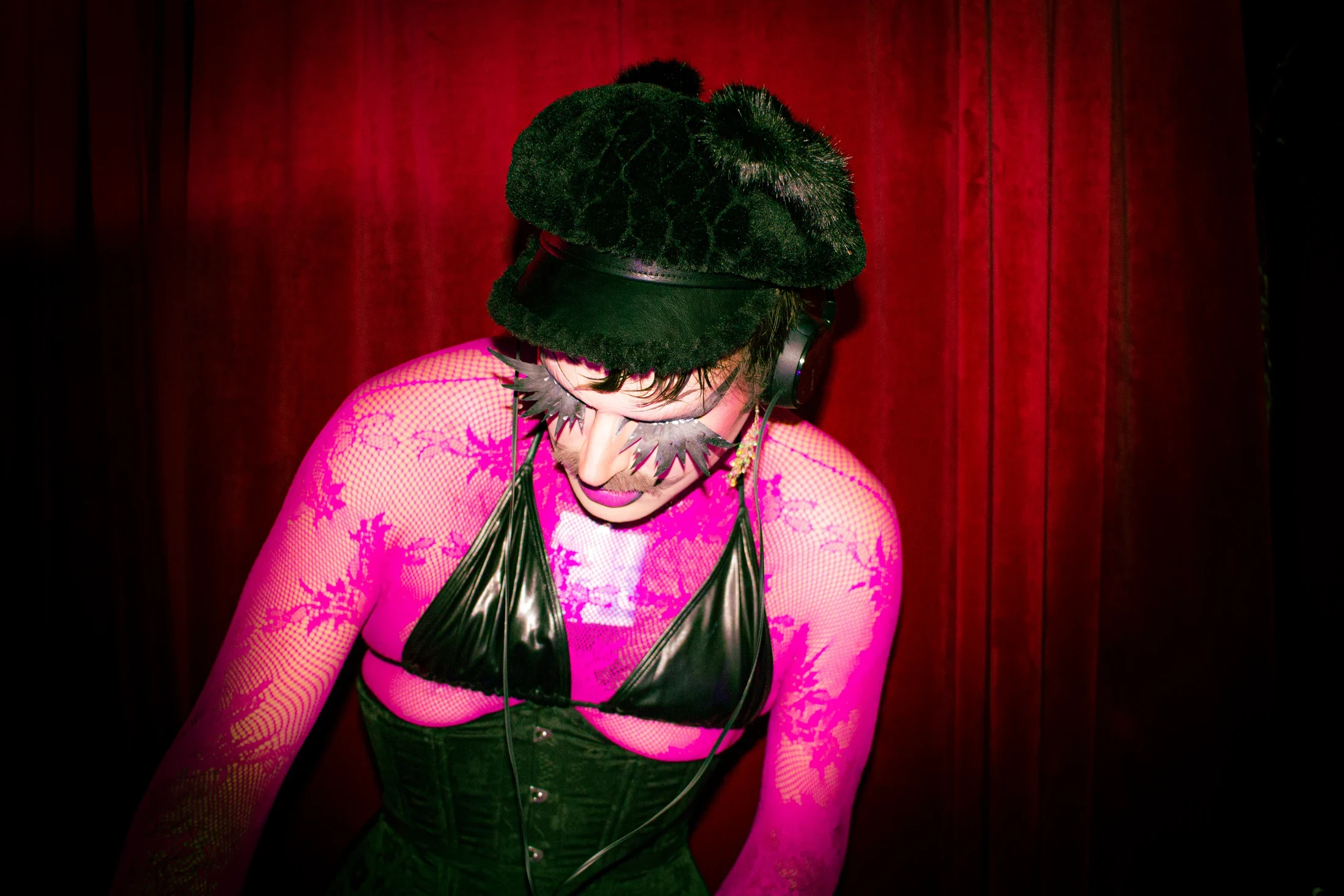 A performer wearing headphones and dramatic star-shaped lashes looks down against a red curtain.
