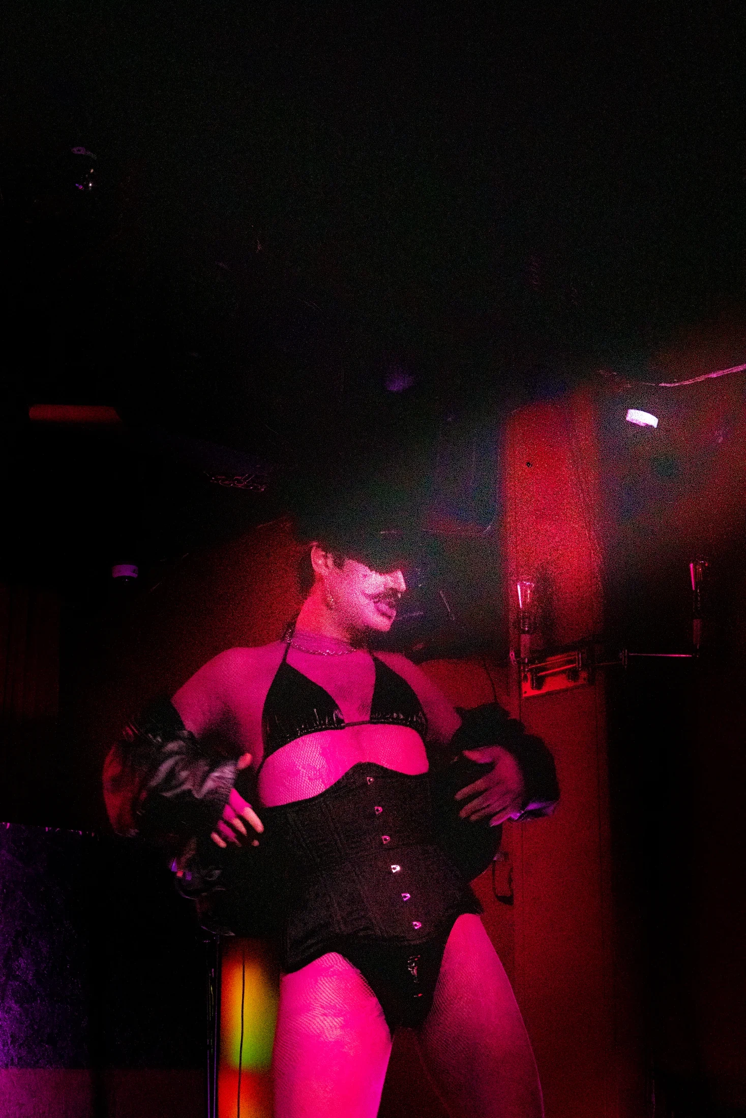 A performer in a black corset poses in a dark room washed with magenta light.