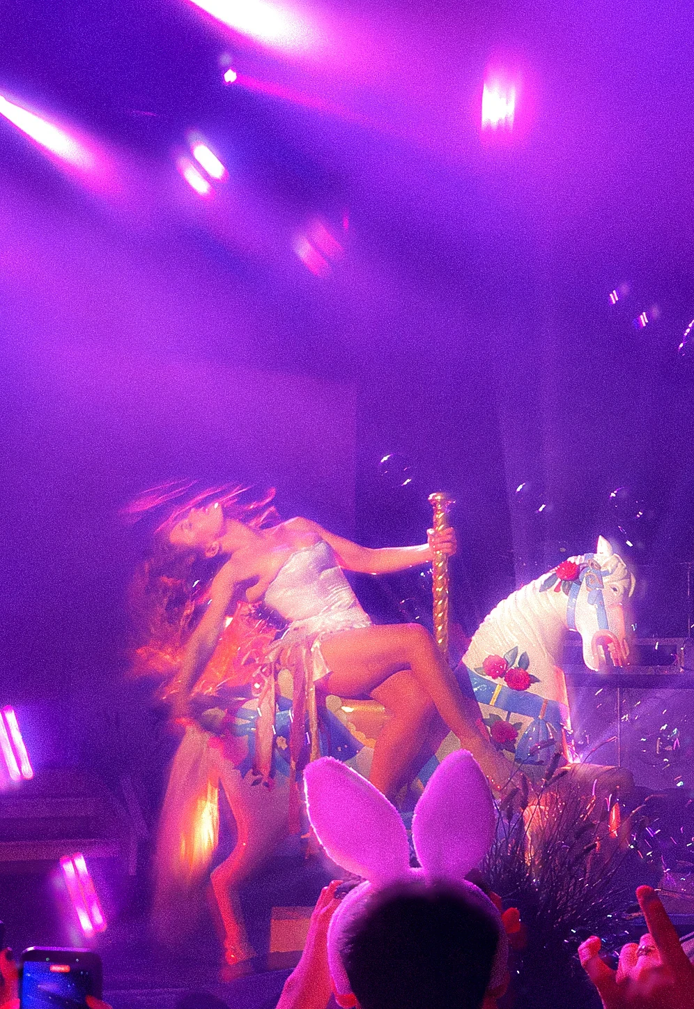 Slayyter leans back on a carousel horse under purple stage lights, framed by the audience.