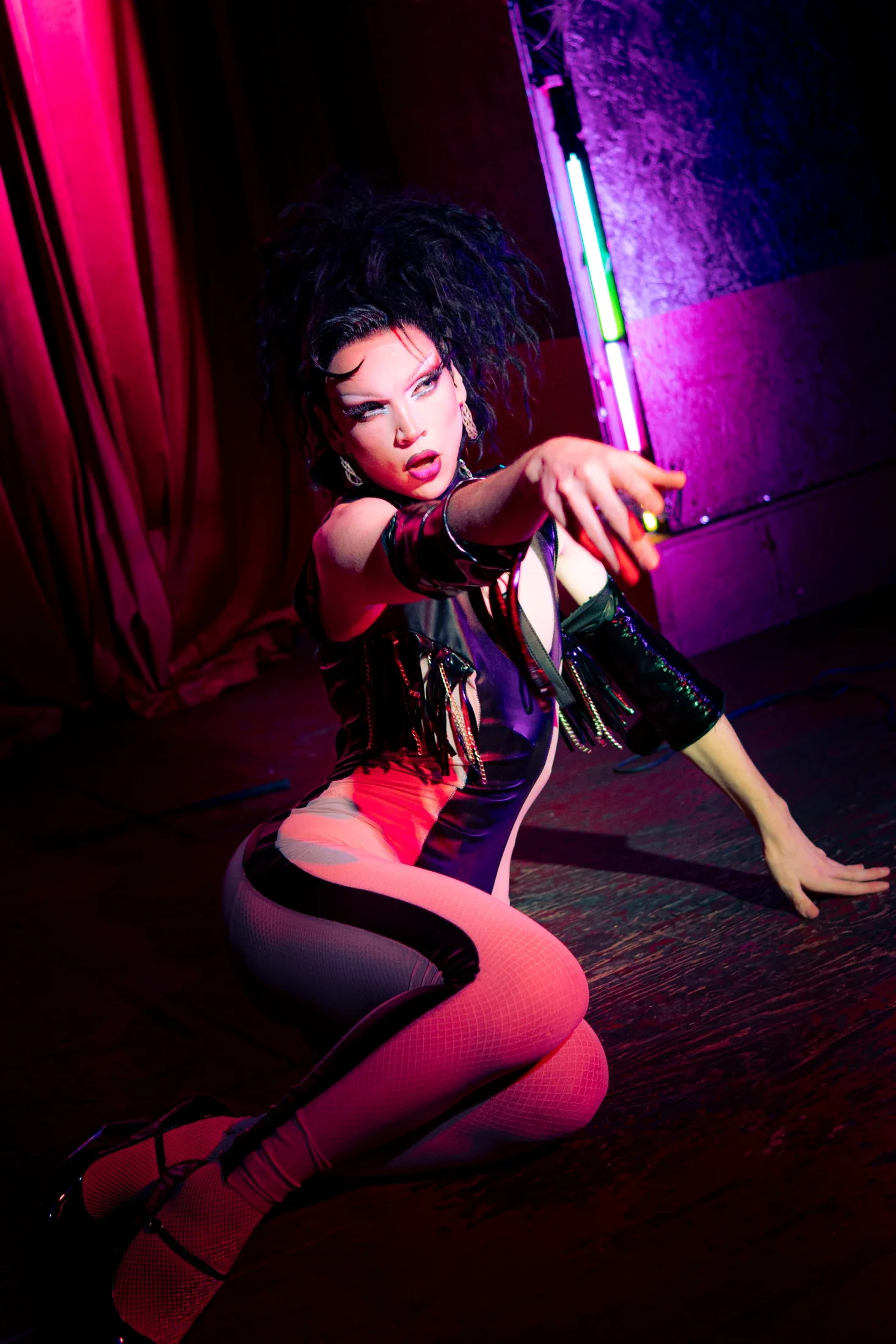 A performer seated on the stage reaches toward the camera under vivid pink and purple lights.