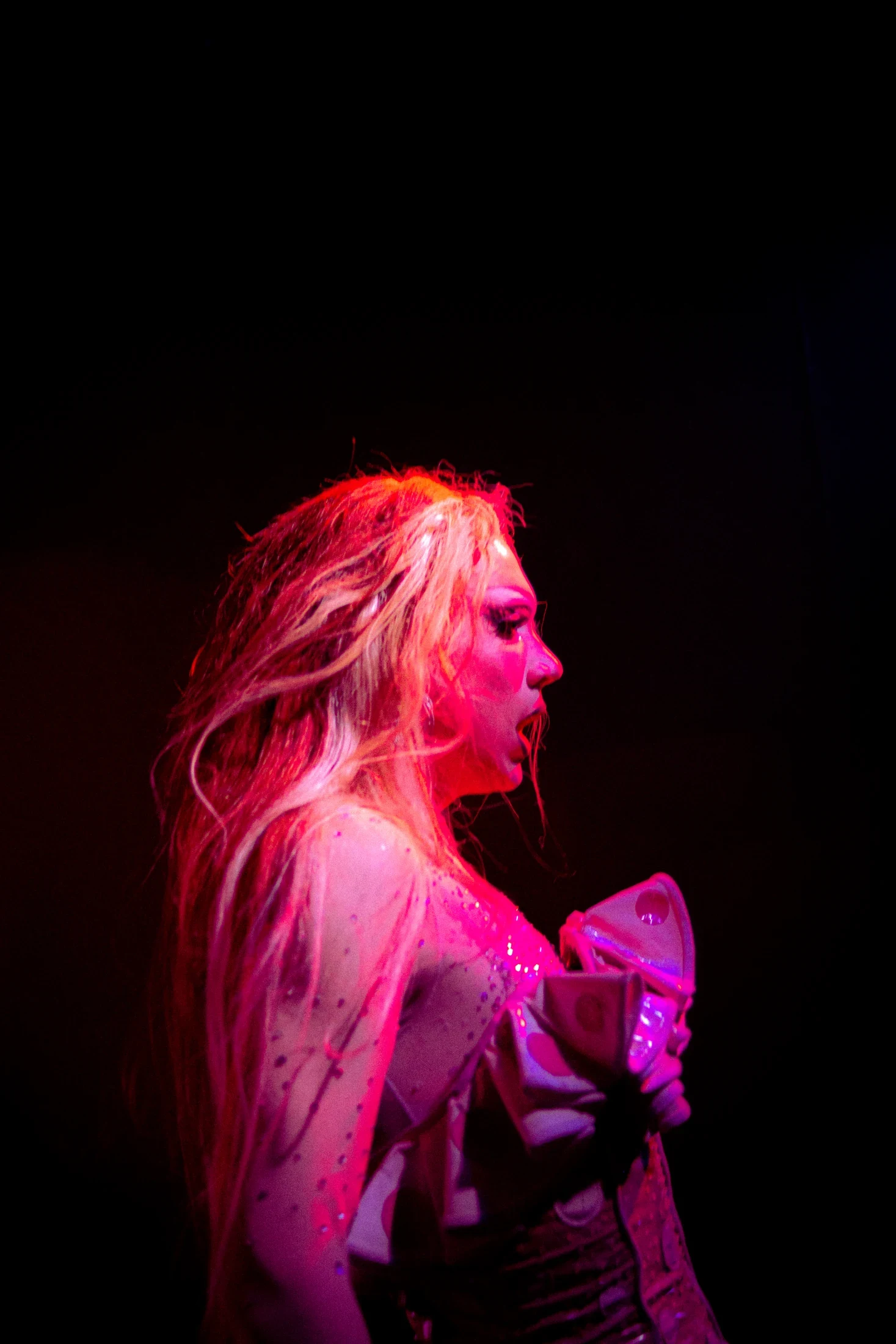 A performer in profile holds patterned fabric against a dark background in pink stage light.